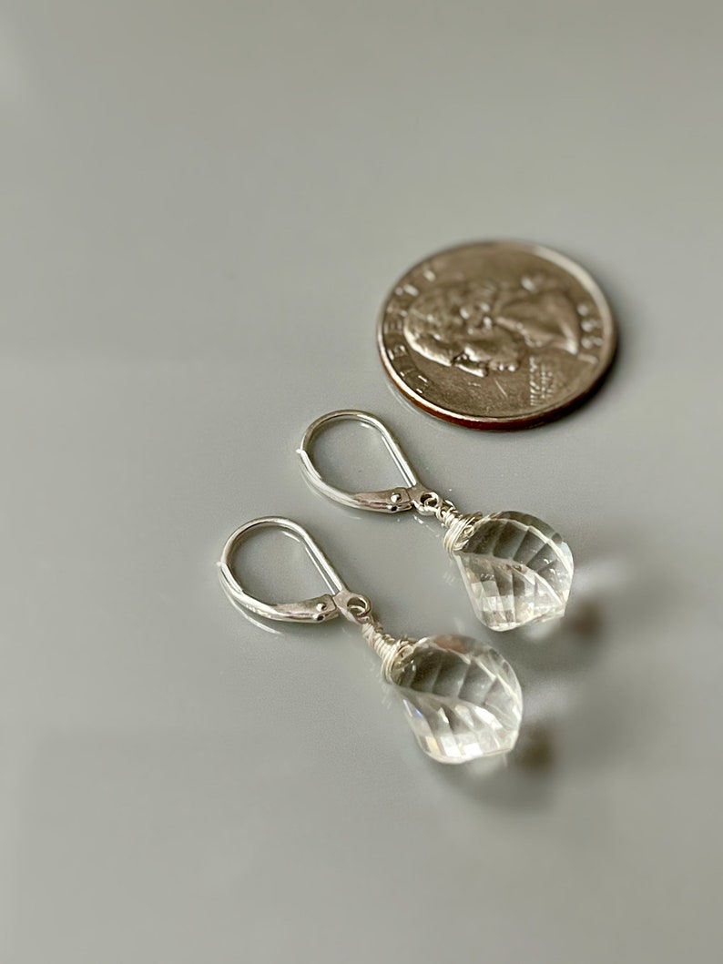 May include: A pair of silver earrings with clear crystal teardrop-shaped stones. The earrings are approximately the size of a US quarter.