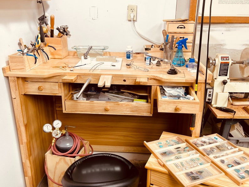 May include: A light-colored wooden jeweler's workbench featuring a central cutout, multiple drawers, and a top surface organized with various metal tools, pliers, a soldering station, a blue spray bottle, and a small wooden organizer box.