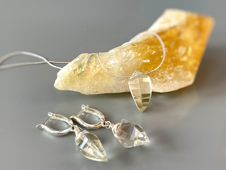 May include: A silver necklace and earrings set with clear quartz crystal pendants. The necklace is on a clear cord and the earrings have silver hoops.