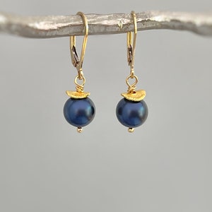 Puede incluir: Un par de pendientes de oro con cierre de palanca y perlas redondas de color azul oscuro. Cada perla est&aacute; rematada con una peque&ntilde;a pieza dorada, texturizada y semicircular, unida al gancho mediante un fino aro y alambre de oro.
