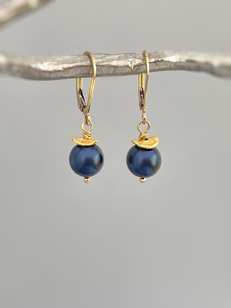 Puede incluir: Un par de pendientes de oro con cierre de palanca y perlas redondas de color azul oscuro. Cada perla est&aacute; rematada con una peque&ntilde;a pieza dorada, texturizada y semicircular, unida al gancho mediante un fino aro y alambre de oro.