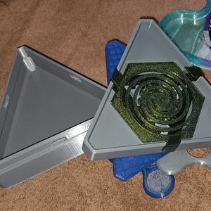 May include: A gray triangular box with a matching lid. The box contains a hexagonal design with a spiral pattern in gold and black. Additional blue and silver shapes are visible.