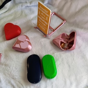 May include: A collection of small, colorful objects. Includes a red heart-shaped object, a pink heart-shaped object, a pink folding object, a navy blue oval object, and a green oval object. A small sign with text is also present.