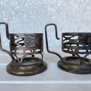 May include: Two antique silver-toned metal tea cup holders. Each has a decorative openwork design with a handle. The holders have a round base and are likely from the early 20th century. The holders are approximately 3 inches tall.