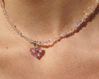 Pode incluir: Um delicado colar de corrente prateada com um pingente de coração de cristal rosa. O colar é feito de pequenos cristais iridescentes.