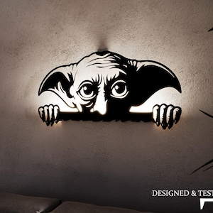 Könnte beinhalten: Eine schwarze Wandskulptur aus Metall, die eine Kreatur mit großen Augen darstellt, die über einen Sims schaut. Die Skulptur wird von hinten beleuchtet, wodurch ein leuchtender Effekt entsteht. Der Text "DESIGNED & TESTED BY T$E" ist in der rechten unteren Ecke des Bildes sichtbar.