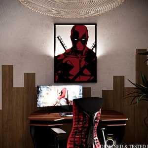 May include: A red and black gaming chair with a mesh back and a black computer desk with a monitor and a framed image of Deadpool. The desk is in a room with wood paneling on the walls.