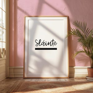 Può includere: Una stampa incorniciata con sfondo bianco e una linea nera sotto la parola "Sláinte" in una cornice dorata.