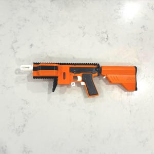May include: A toy blaster featuring a bright orange body with black accents on the grip, top rail, and barrel shroud. The design includes a white muzzle tip, a white trigger, and a solid orange stock. The construction appears to be 3D printed.