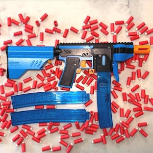 May include: A blue and black toy blaster with an orange tip, featuring a long blue translucent magazine inserted and two additional matching magazines. The blaster is surrounded by numerous small red foam darts with white tips.