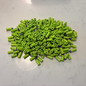 May include: A pile of bright green foam darts with purple tips. The darts are cylindrical and appear to be for a toy gun. The darts are scattered on a white surface.