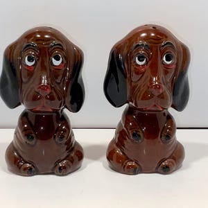 May include: A pair of ceramic dachshund dog figurines. The salt and pepper shakers are brown with black ears and white-rimmed eyes. The dog figurines have a glossy finish and are sitting upright.