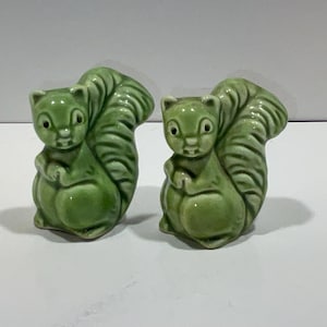 May include: Two ceramic squirrel figurines in a glossy green finish. The squirrels are depicted in a sitting position with their bushy tails arched over their backs. These decorative figurines are suitable for home decor.