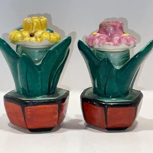May include: A pair of ceramic salt and pepper shakers. Each shaker is shaped like a flower in a pot. One flower is yellow, the other is pink. The pots are orange with black trim, and the leaves are green.
