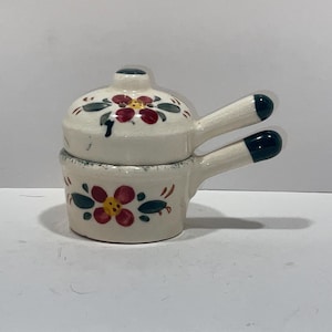 May include: A small, white ceramic pot with a lid, featuring red and green floral designs. The pot has two handles and a matching lid with a small knob. The handles and knob have dark green tips. A decorative kitchen item.