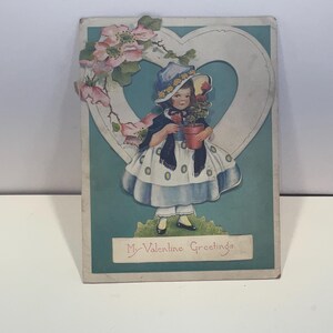 May include: Vintage Valentine's Day postcard featuring a young girl holding a potted plant. The girl is dressed in a blue and white dress and hat, standing in front of a heart-shaped frame. Pink flowers and the text "My Valentine Greetings" are also visible.