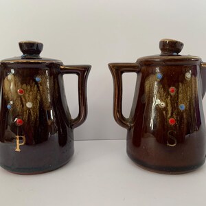 May include: A set of two brown ceramic salt and pepper shakers with a speckled glaze. The shakers are shaped like small teapots with a handle and spout. The shakers are decorated with white, red, and blue dots. The word "P" is written on one shaker and the word "S" is written on the other.