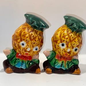 May include: Two ceramic pineapple-shaped figurines. Each figurine has a yellow and brown pineapple body, green hat, and a red bow tie. The figurines have blue eyes, and are wearing green jackets and brown pants.