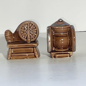 May include: A pair of brown ceramic figurines. One is a spinning wheel, the other a lantern. The spinning wheel has a detailed wheel design. The lantern has a silver-colored trim. Both are on a white surface.