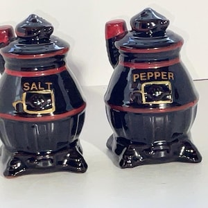 May include: A pair of vintage black ceramic salt and pepper shakers. Each shaker has a rounded body with a red band and a handle. The word "SALT" is on one shaker and "PEPPER" on the other, both in gold lettering.