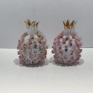 May include: Two decorative pineapple-shaped ornaments. Each ornament is white with pink rose accents and gold-colored leaves. The pineapples are ceramic and sit on a white surface.