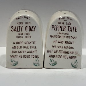 May include: A pair of novelty salt and pepper shakers shaped like tombstones. Each shaker is off-white with brown text, featuring humorous epitaphs and dates. Green floral accents are at the bottom.