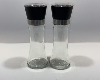 May include: Two clear glass salt or pepper grinders with black tops and stainless steel bands. The grinders have a classic hourglass shape and are designed for kitchen use. The clear glass allows for easy viewing of the contents.