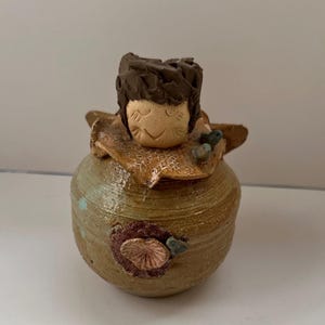 May include: A ceramic figurine of a person with a smiling face. The figurine is decorated with brown, tan, and blue accents. The figurine is sitting on a white surface.