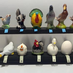 May include: A collection of ceramic bird figurines, including ducks, chickens, and a turkey, displayed on a black tiered shelf. The figurines are painted in various colors and styles, showcasing a range of avian designs.
