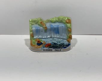 May include: A ceramic Niagara Falls souvenir figurine. The miniature waterfall scene features blue water cascading over a cliff, with green foliage and orange accents. The base is labeled "NIAGARA FALLS".