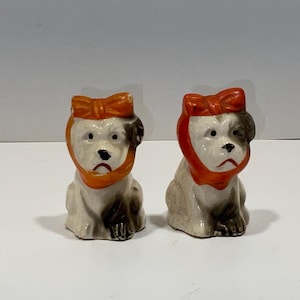 May include: Two ceramic dog figurines. Each dog is white with brown patches, a black nose, and an orange bow on its head and around its neck. The dogs are sitting upright and have a slightly sad expression.