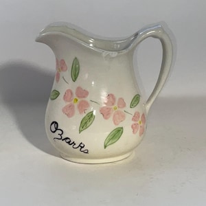 May include: A small, off-white ceramic pitcher with a handle and spout. The pitcher is decorated with pink floral designs and green leaves. The word "Ozarks" is written in black script on the side.