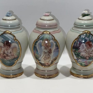 May include: Three decorative ceramic jars with lids, each featuring a different angel illustration within a gold-trimmed oval frame. The jars have a light blue, pink, and green color scheme, with floral accents on the lids and gold trim around the base.