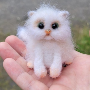 Puede incluir: Una pequeña figura de gato hecha a mano con fieltro de aguja de lana blanca esponjosa. El gato presenta ojos oscuros y redondos, una pequeña nariz naranja y un pelaje con sutiles tonos anaranjados en las orejas y las patas.