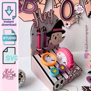 Puede incluir: Un puesto de manualidades de Halloween rosa y negro con una bruja, un fantasma y una calavera. El puesto tiene un patrón a rayas e incluye un pequeño anillo naranja, una cinta métrica rosa y tres lápices. El puesto dice "Hey" y "Boo" en él.
