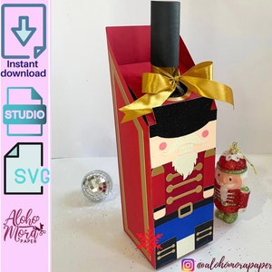 Puede incluir: Un porta botellas de vino de cartón rojo y dorado con forma de cascanueces. El porta botellas tiene un lazo dorado y un pequeño adorno de cascanueces.