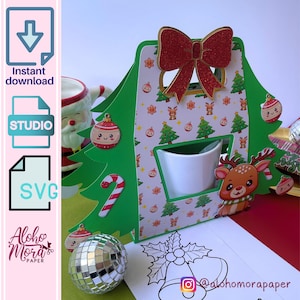 Puede incluir: Una caja verde en forma de árbol de Navidad con un portavasos blanco y un lazo rojo en la parte superior. La caja está decorada con un patrón blanco y rojo de árboles de Navidad, renos y bastones de caramelo. La caja está sobre una superficie blanca con un contorno blanco y negro de una campana.