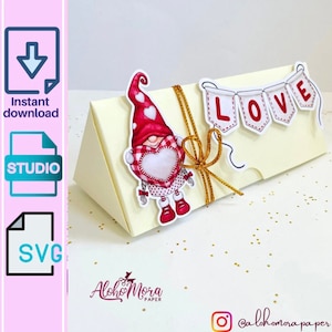 May include: A white triangular box with a red and white gnome holding a heart. The box has a gold ribbon and a banner that says "LOVE" in red letters.