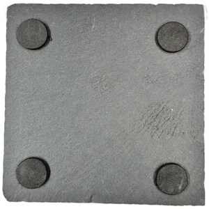 May include: A square, gray slate coaster with four round, dark gray pads on the corners. The coaster has a textured surface and is designed to protect surfaces from drink condensation.