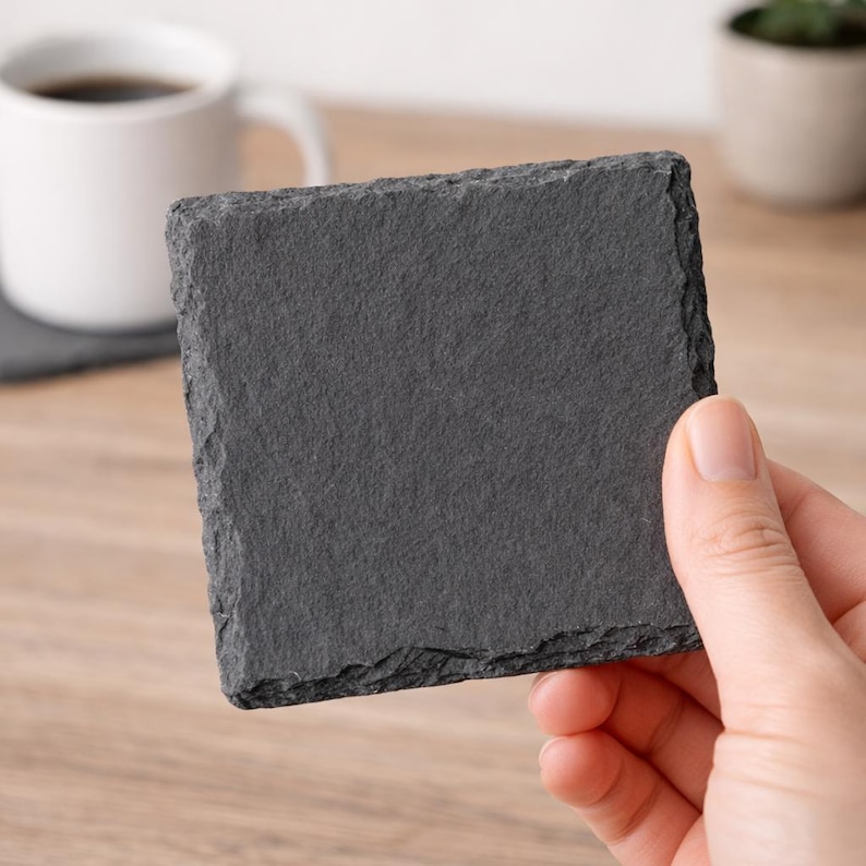 May include: A square, dark gray slate coaster with a rough, textured surface. The coaster is held by a hand, showcasing its natural, uneven edges. A white coffee mug and a small plant are visible in the background, suggesting a home decor theme.