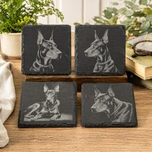 May include: Set of four square black slate coasters. Each coaster features a detailed engraved portrait of a Doberman Pinscher dog. The dog portraits are in profile, with varying poses, and are set against a dark background.