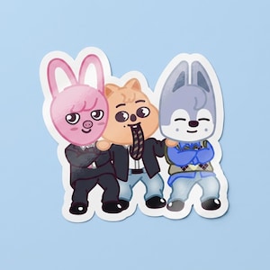 Op de afbeelding: Een sticker met drie stripfiguren: een roze konijn, een bruine hamster en een grijze wolf. Ze dragen pakken en truien, met de tong van de hamster uitgestoken. De sticker heeft een witte rand en een lichtblauwe achtergrond.
