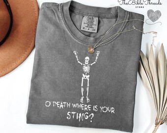 May include: A gray Comfort Colors t-shirt with a white skeleton graphic and the text "O DEATH WHERE IS YOUR STING?". A silver necklace with a pendant is on the shirt. The Bible Threads store logo is in the upper right corner.