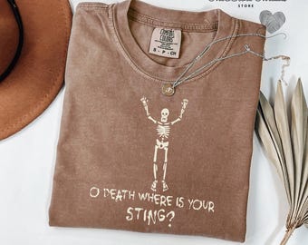 May include: A brown Comfort Colors t-shirt with a skeleton graphic and the text "O DEATH WHERE IS YOUR STING?". The shirt also features the text "The Bible Threads STORE". A silver necklace is draped across the shirt.