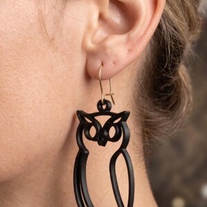 May include: Black owl-shaped earrings with gold-coloured hooks. The earrings are a cutout design, showing the outline of an owl. The owl's eyes, beak, and feet are visible. The earrings are worn on a person's ear.