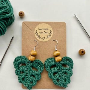 May include: Handmade green crochet leaf earrings with wooden beads and silver hooks. The earrings are displayed on a small brown card with a sticker that says "handmade with love". A ball of green yarn, a needle, and a crochet hook are also in the image.