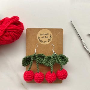 Op de afbeelding: Gehaakte kersenoorringen met groene bladeren en zilveren haakjes. De oorbellen worden gepresenteerd op een bruine kaart met de tekst "handmade with love".