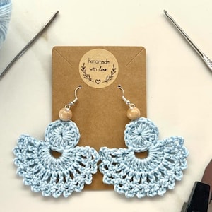 Może przedstawiać: Para niebieskich, szydełkowych kolczyków z drewnianymi koralikami i srebrnymi haczykami. Kolczyki są prezentowane na brązowej kartce z napisem "ręcznie robione z miłością".