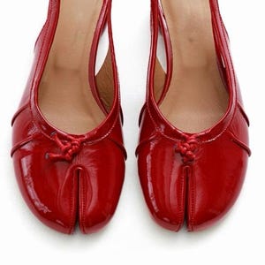 May include: Pair of red patent leather slingback flats. These shoes feature a split-toe design, a small knot detail on the vamp, and adjustable ankle straps. The interior is a light tan color, and the shoes are set against a white background.