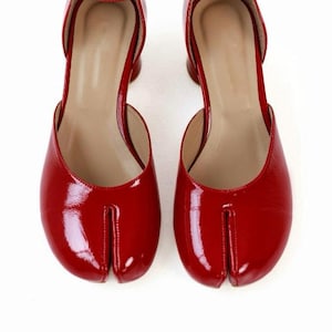 May include: Pair of red patent leather Tabi shoes with ankle straps and small heels. The shoes have a split-toe design, a signature feature of Tabi footwear, and a glossy finish. The shoes are set against a white background.